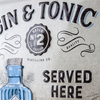 Decorative sign with 'Gin & Tonic Served Here' text and a blue bottle illustration on a metallic background.