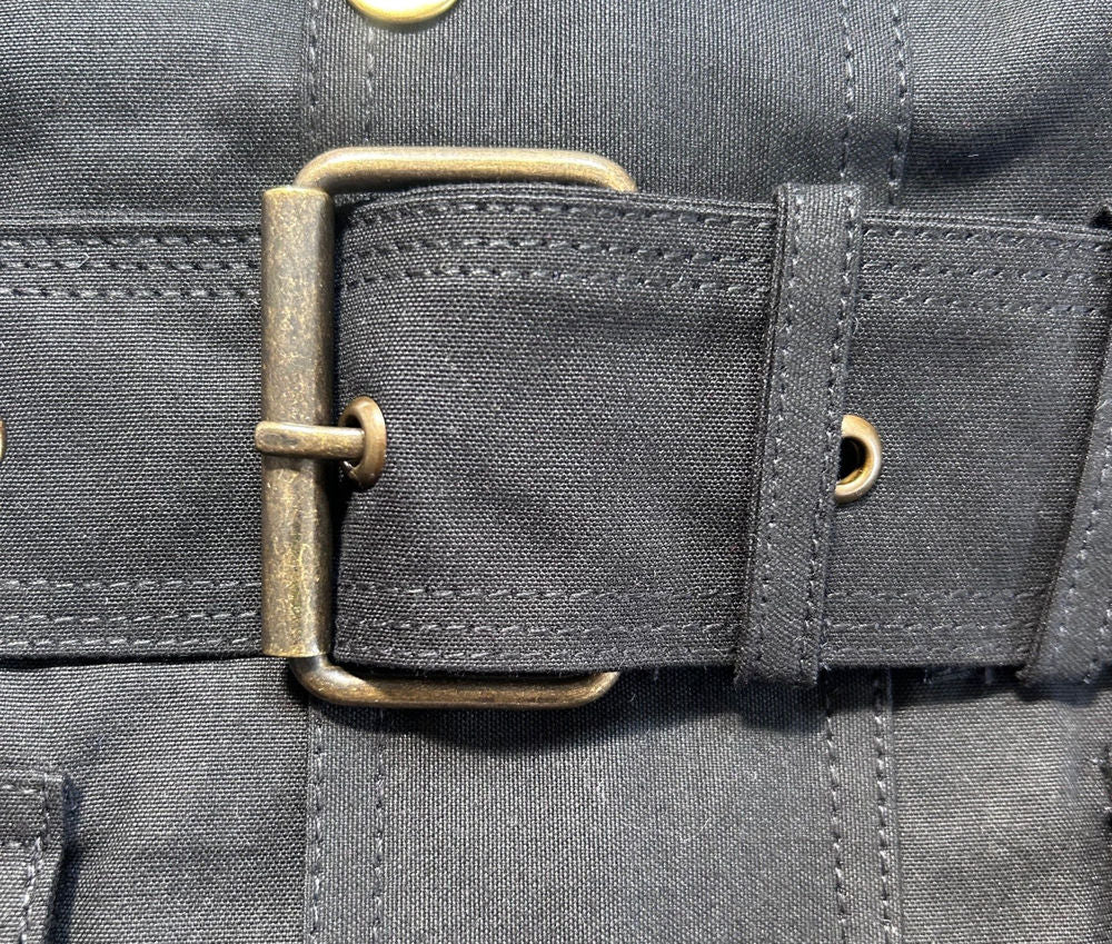 Close-up of a black waxed cotton jacket belt with a brass roller buckle and eyelets on a matching black garment.